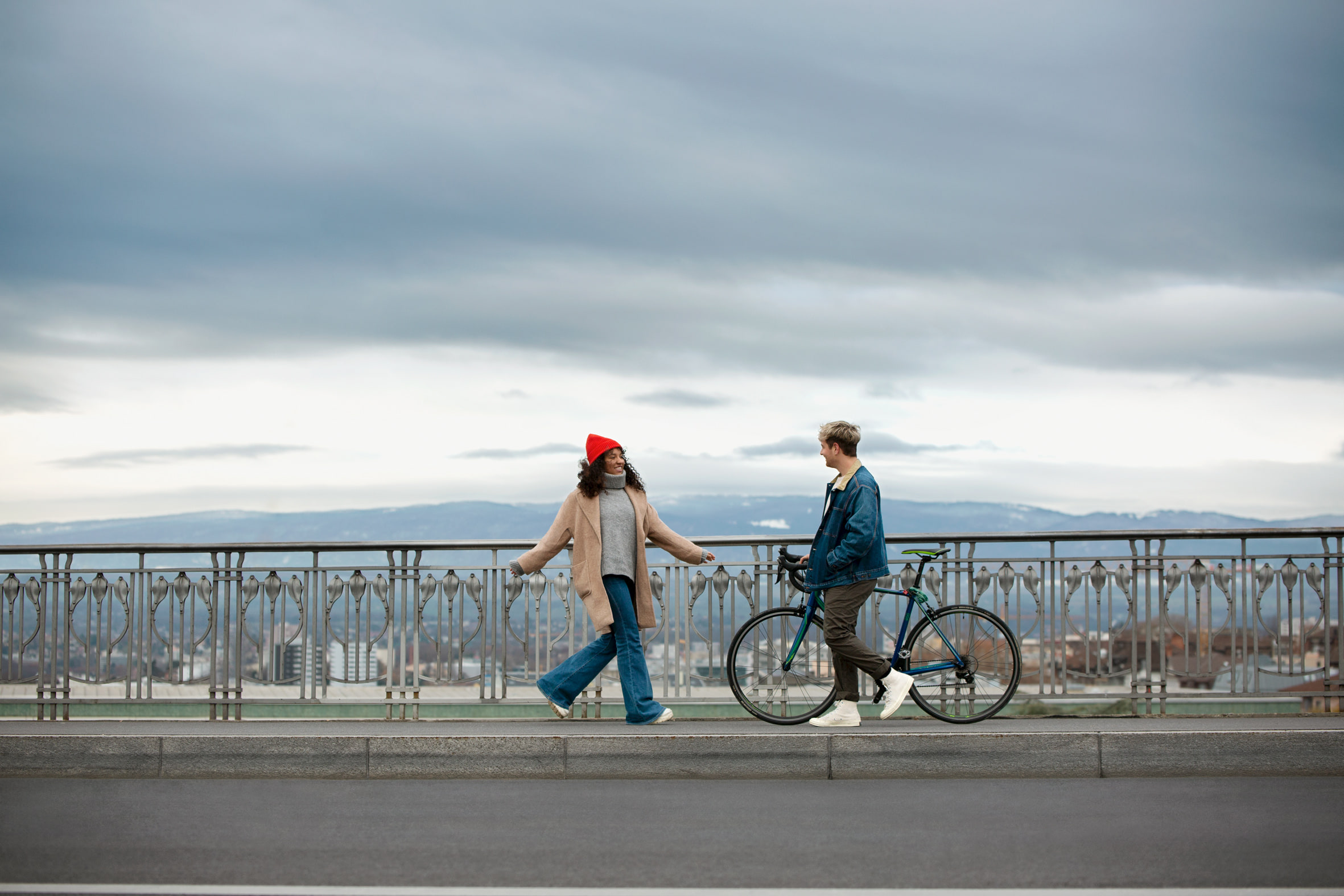 Femme au bonnet rouge et homme poussant un vélo qui se croisent sur un pont, montagnes en arrière-plan
