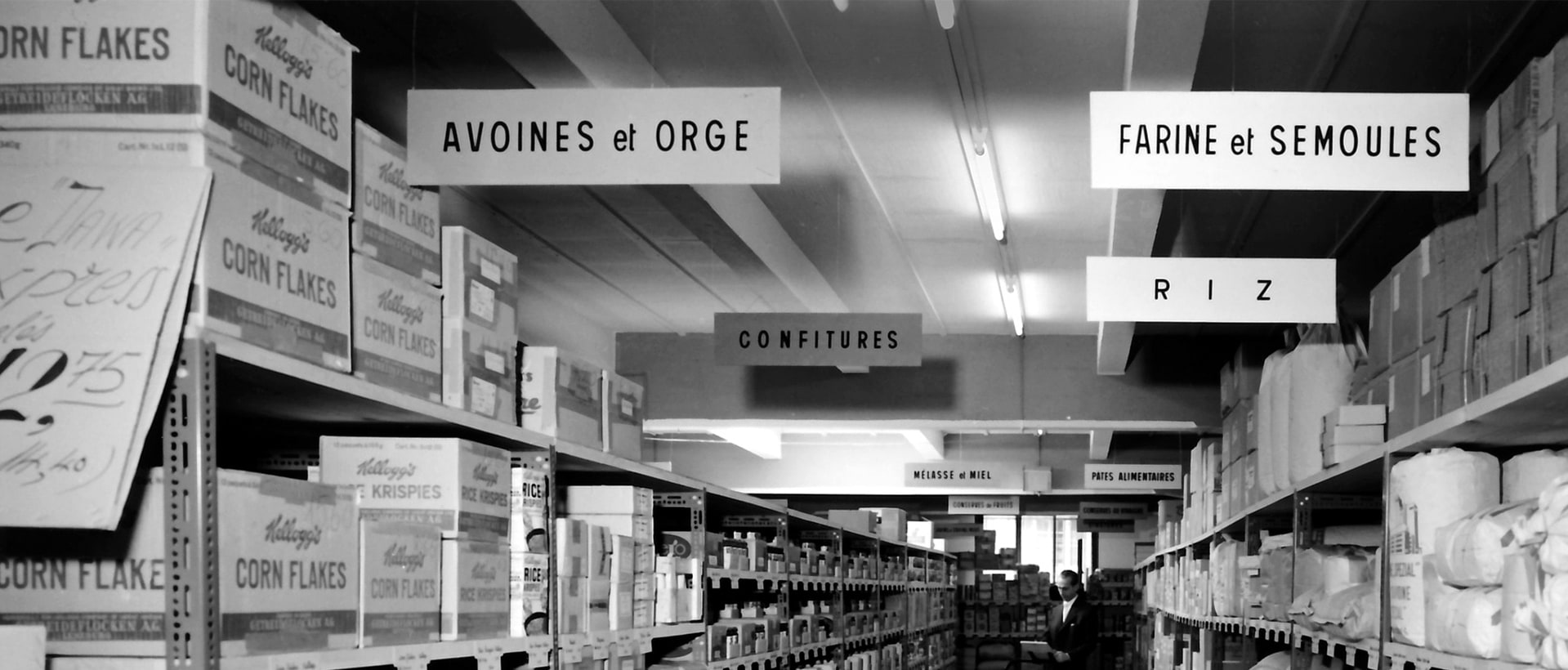 Allées d’un ancien entrepôt en noir et blanc, rayons de corn flakes et panneaux « Avoines et orge », « Farine et semoules », « Riz »