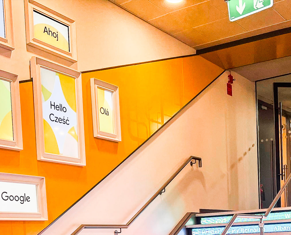 Une cage d’escalier orange, des cadres au mur disant bonjour en plusieurs langues : Ahoj, Hello, Cześć, Olá.