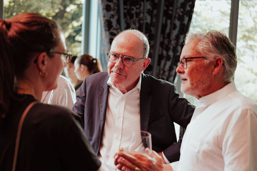 Deux participants échangent, un verre à la main, devant les rideaux de la salle.