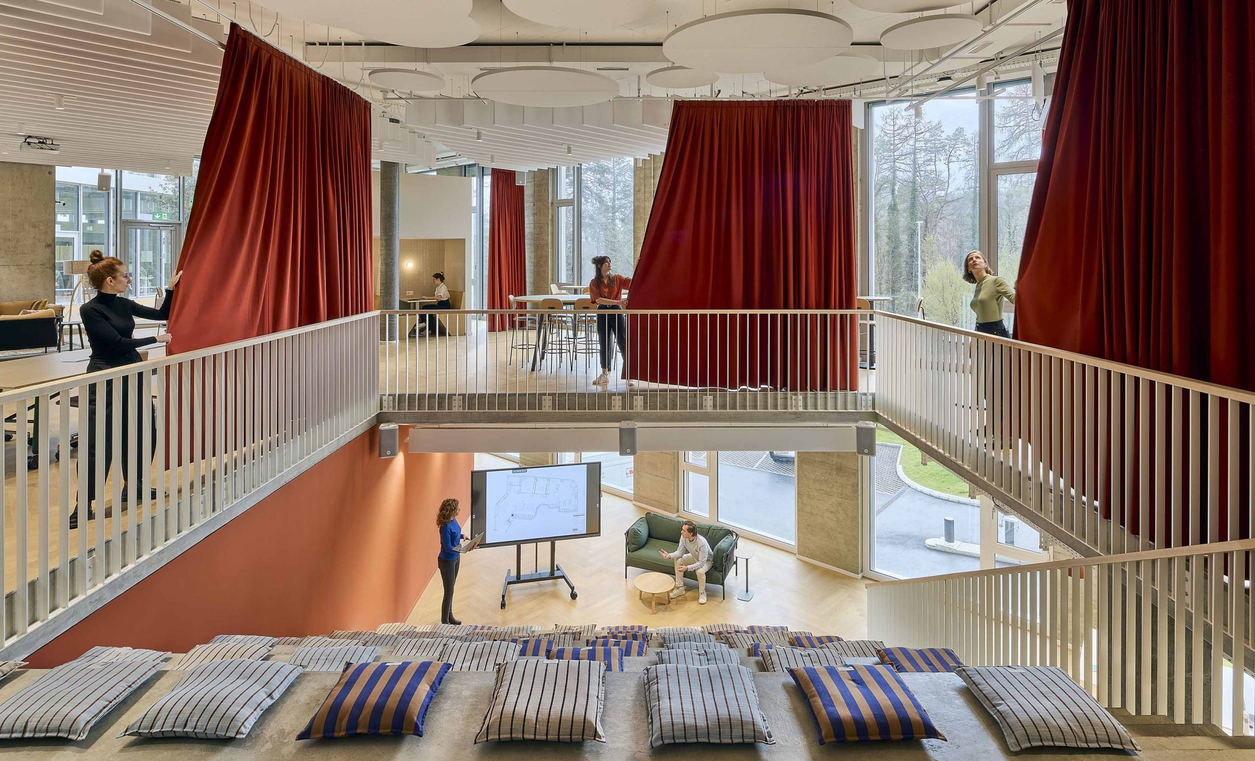 Espace de travail ouvert sur deux niveaux, avec rideaux rouges, gradins à coussins et écran de présentation