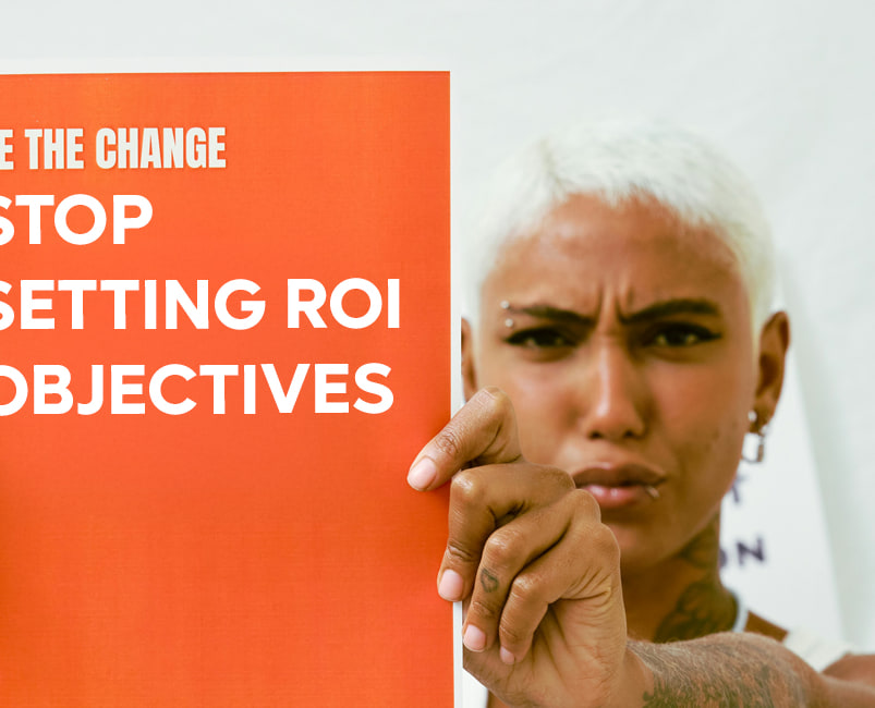 Une femme brandit une pancarte orange disant « Be the change — stop setting ROI objectives », et pointe l’objectif du doigt.