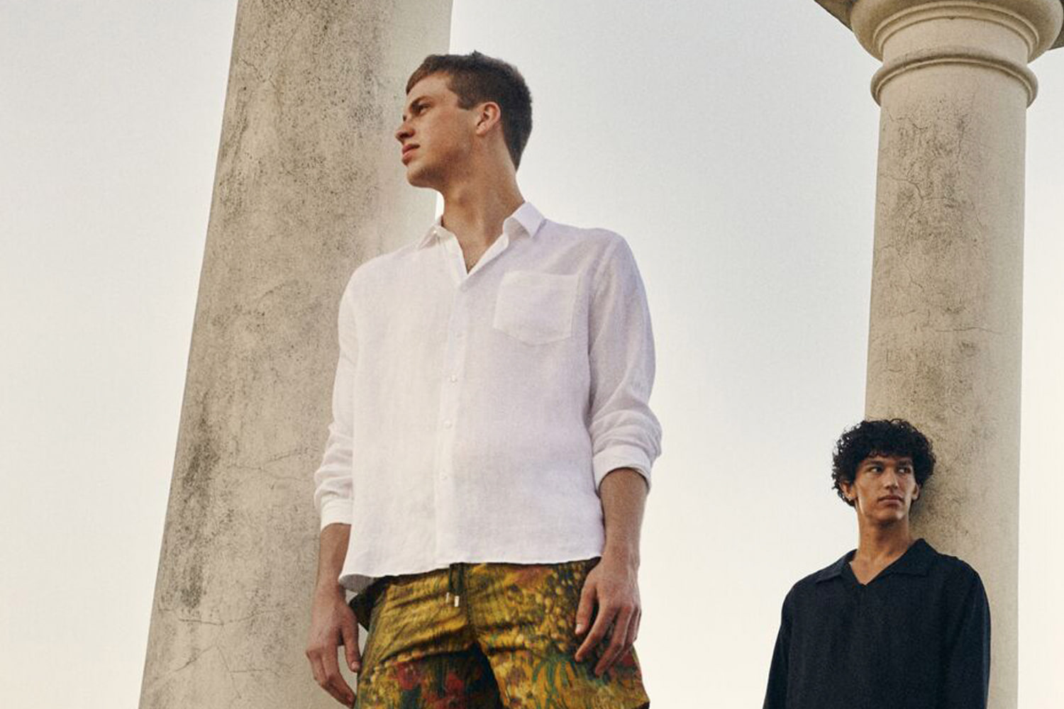 Deux jeunes hommes en chemise entre des colonnes antiques, l’un en chemise de lin blanche et short imprimé